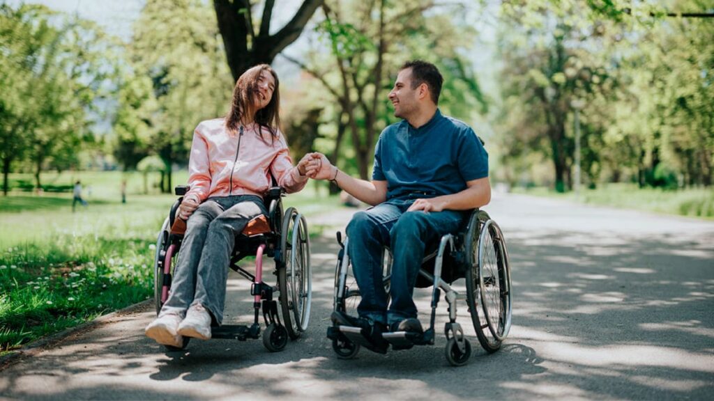 Um casal de usuários de cadeira de rodas, um homem e uma mulher, passeiam de mãos dadas em um parque arborizado, sorrindo um para o outro. (Foto: Depositphotos)
