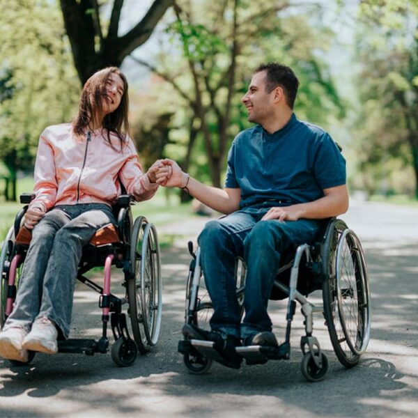 Um casal de usuários de cadeira de rodas, um homem e uma mulher, passeiam de mãos dadas em um parque arborizado, sorrindo um para o outro. (Foto: Depositphotos)