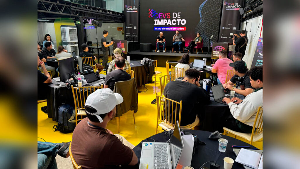 Participantes do evento 'Devs de Impacto' assistem a um painel de debate sobre Inteligência Artificial, com desenvolvedores em mesas redondas usando seus laptops. (Foto: Divulgação)