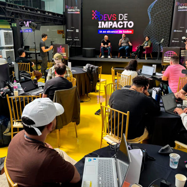 Participantes do evento 'Devs de Impacto' assistem a um painel de debate sobre Inteligência Artificial, com desenvolvedores em mesas redondas usando seus laptops. (Foto: Divulgação)