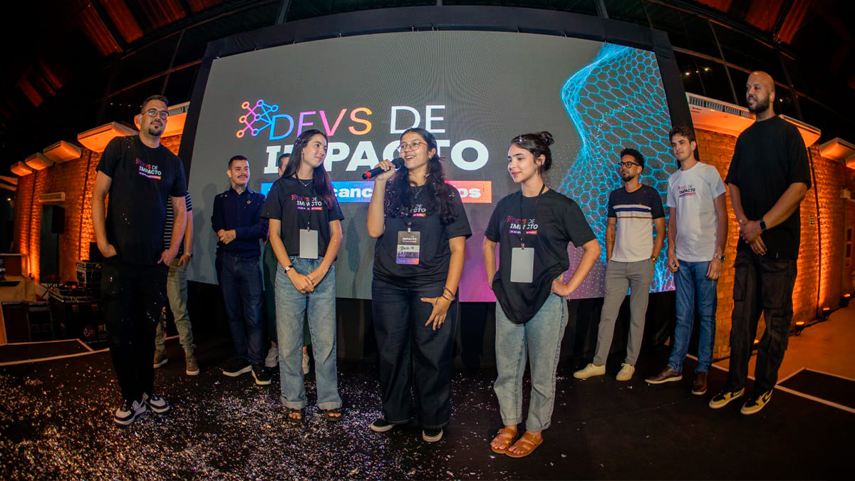 Equipe vencedora do hackathon Devs de Impacto, composta por três mulheres, no palco durante a premiação, com uma delas falando ao microfone. (Foto: Divulgação/iMasters)
