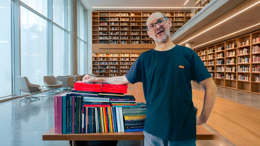O pesquisador Emílio Figueira, sorrindo, ao lado de uma grande pilha de livros em uma biblioteca moderna.