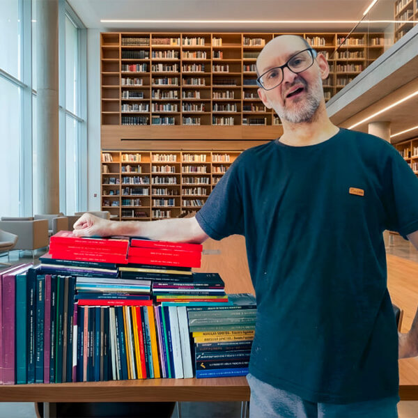 O pesquisador Emílio Figueira, sorrindo, ao lado de uma grande pilha de livros em uma biblioteca moderna.