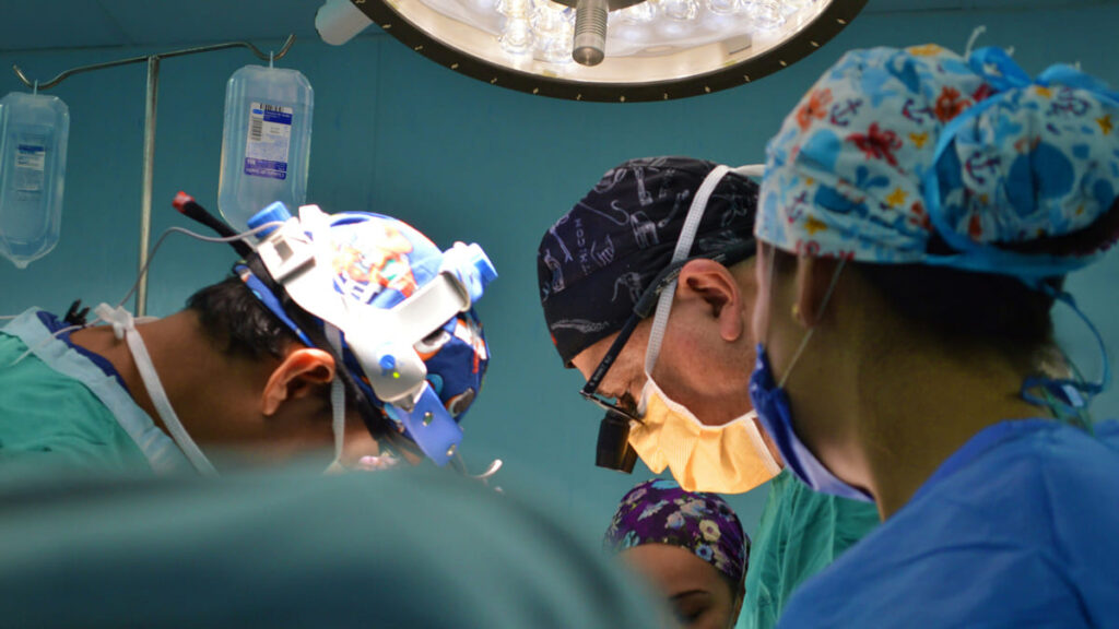 Equipe de cirurgiões usa lupas de precisão durante procedimento de implante neural, focados sob a luz do centro cirúrgico.