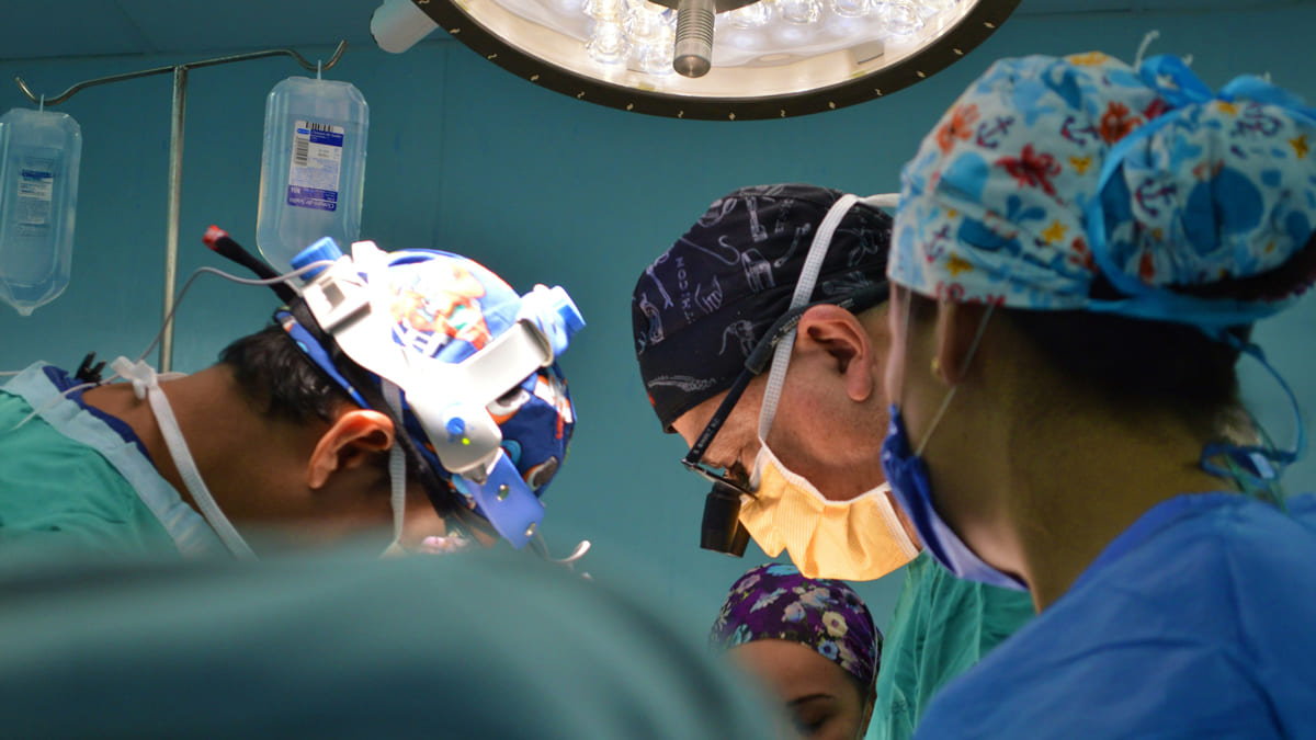 Equipe de cirurgiões usa lupas de precisão durante procedimento de implante neural, focados sob a luz do centro cirúrgico.