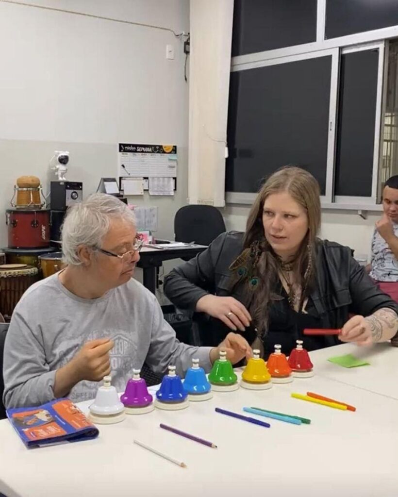 Em uma sala com instrumentos, um idoso e uma terapeuta tocam sinos coloridos de mão alinhados sobre uma mesa branca.