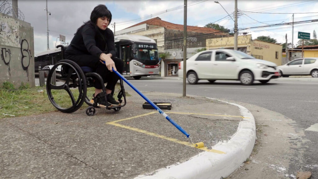 Luiza Sigulem, em cadeira de rodas, usa um rolo de cabo longo para pintar uma demarcação amarela na guia de uma calçada.