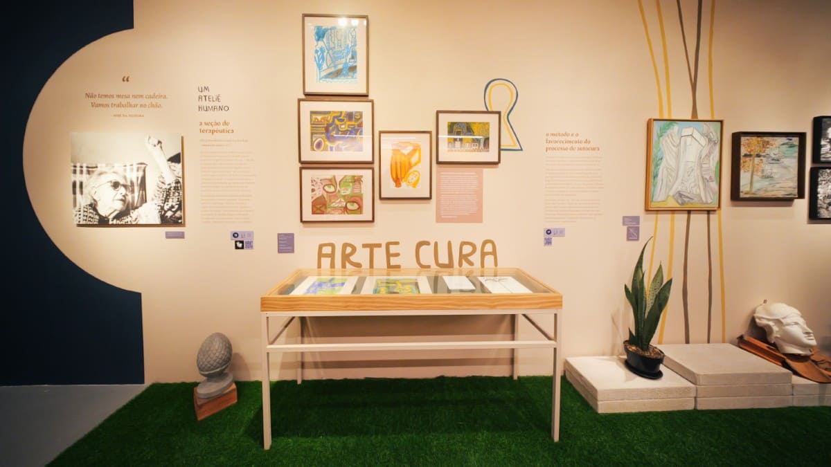 Fotografia da Exposição de Nise com seu retrato, quadros com gravuras e texto em destaque 'Arte Cura'.