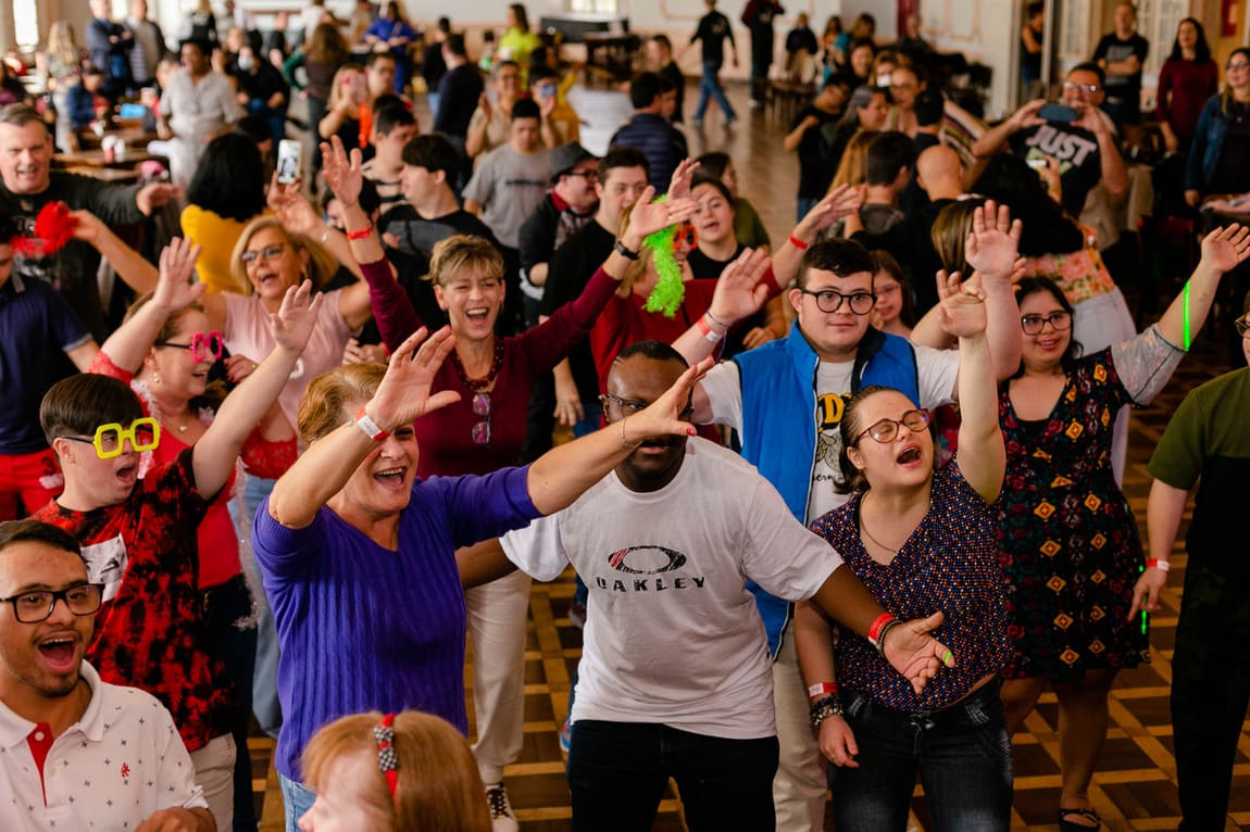 Carnaval Baladown: Grupo diverso de pessoas, incluindo jovens e adultos com Síndrome de Down, dançando com os braços levantados em um salão de festa.