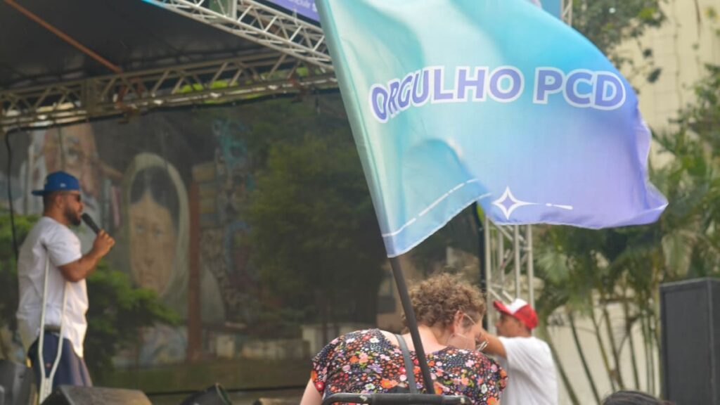 Bandeira azul com o texto "ORGULHO PCD" em destaque. Ao fundo, um homem usando muletas fala ao microfone no palco do evento.