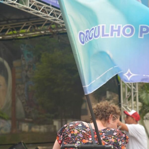 Bandeira azul com o texto "ORGULHO PCD" em destaque. Ao fundo, um homem usando muletas fala ao microfone no palco do evento.