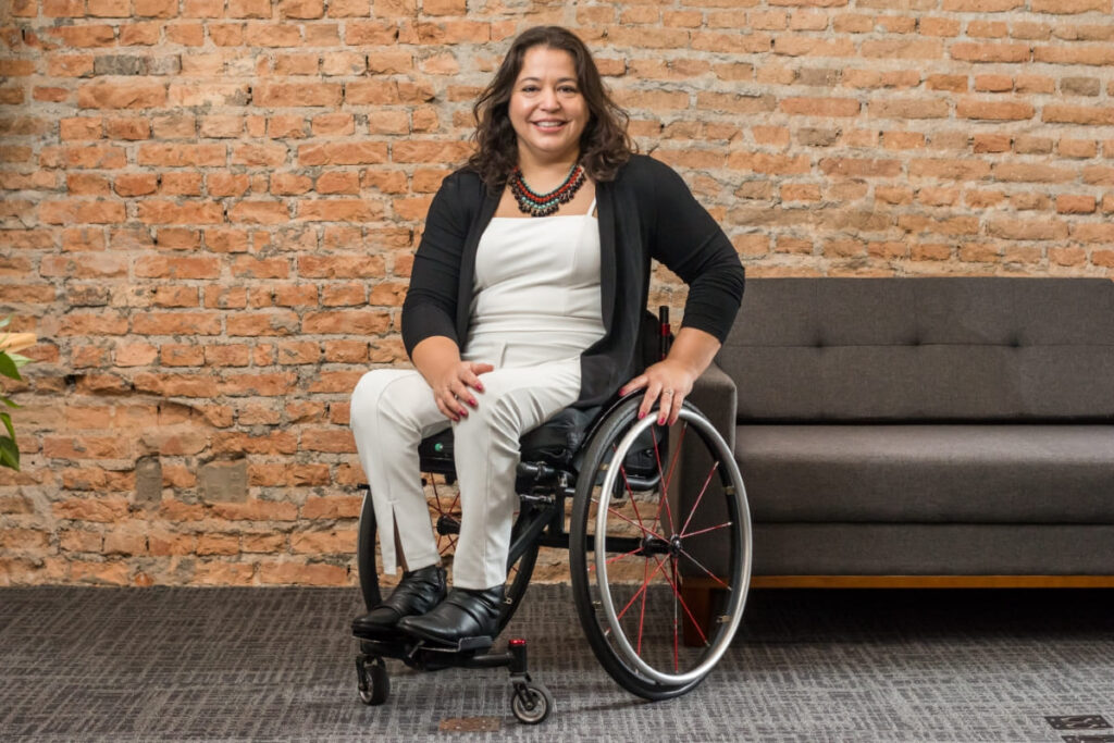Carolina Ignarra, CEO da Talento Incluir, sorri sentada em sua cadeira de rodas manual, em frente a uma parede de tijolos.