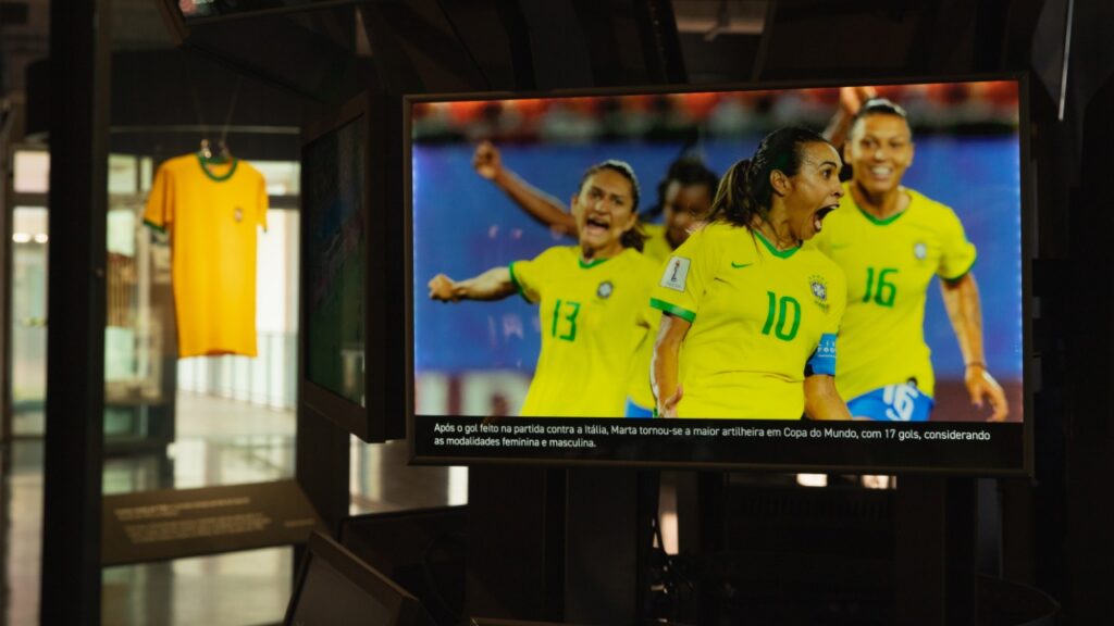 Tela no Museu do Futebol exibe a jogadora Marta (camisa 10) comemorando um gol pela Seleção Brasileira junto à sua equipe