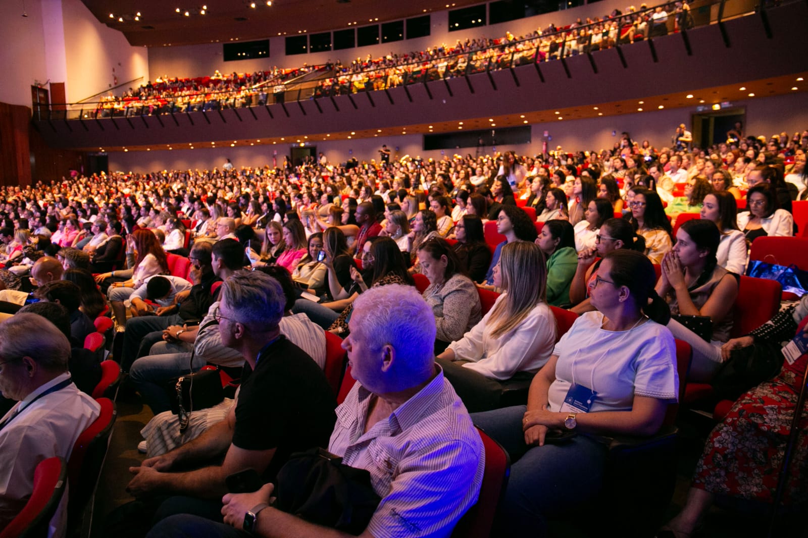 Grande auditório lotado. Centenas de pessoas estão sentadas em poltronas vermelhas, voltadas para o palco do congresso.