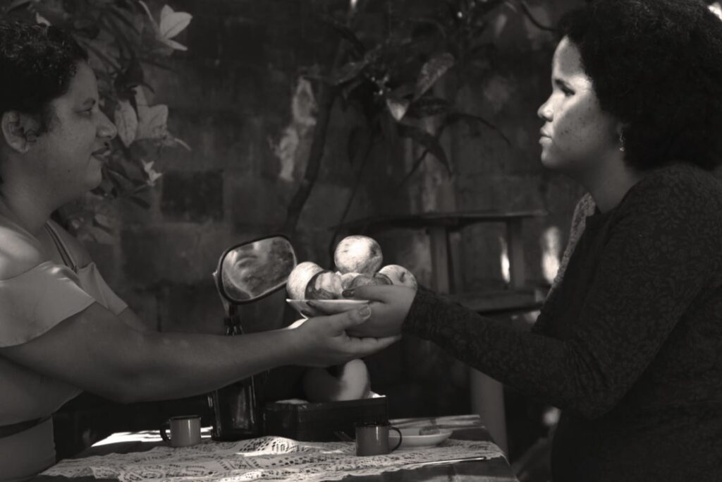 Fotografia em preto e branco de duas mulheres sentadas frente a frente em uma mesa. Uma entrega um prato com maçãs para a outra.