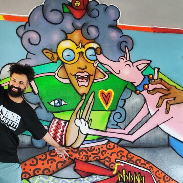 O artista Enivo sorri com o braço erguido ao lado do mural de arte urbana em cores e acessível, no CEU Perus.