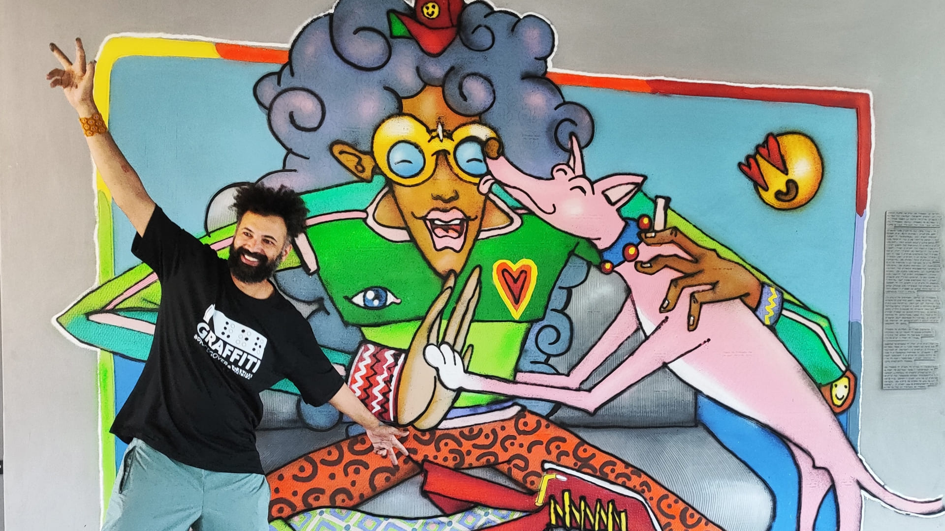 O artista Enivo sorri com o braço erguido ao lado do mural de arte urbana em cores e acessível, no CEU Perus.