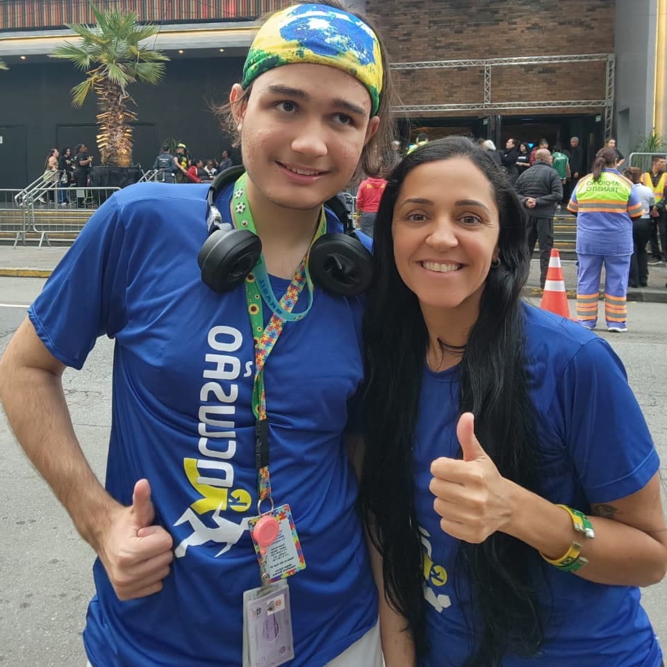 Juliane de Araújo e seu filho Geraldo sorriem e fazem sinal de positivo. Ele usa abafadores de ruído e cordão de girassol.