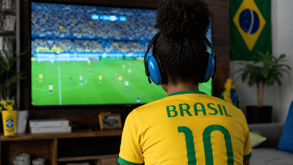 Pessoa com camisa 10 do Brasil usa abafadores de ruído azuis enquanto assiste a um jogo de futebol na TV da sala.