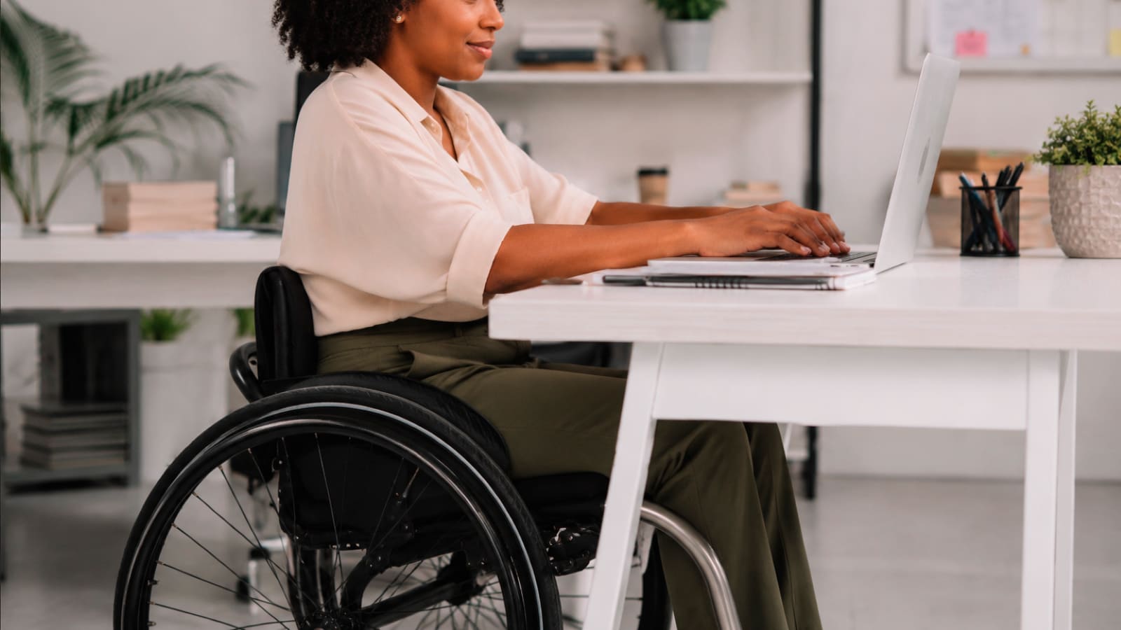 Profissional negra em cadeira de rodas digita em um laptop sobre uma mesa branca em um escritório moderno e iluminado.