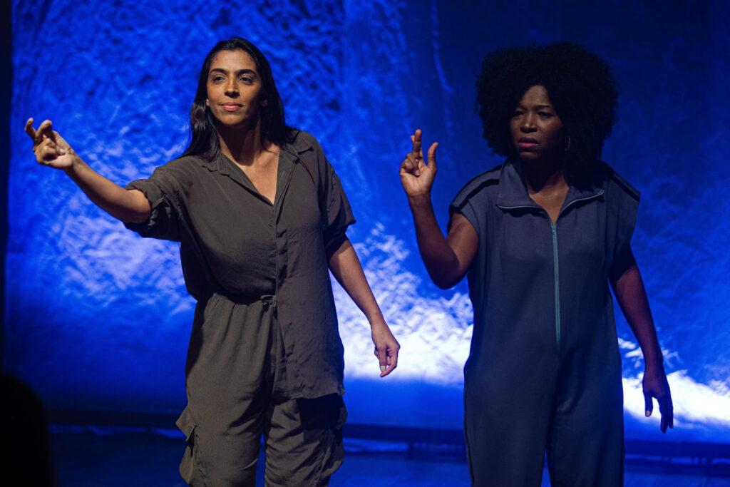Cena da peça SURDA, cultura DEF no festival Acessa BH 2026. Duas atrizes, em pé num palco com iluminação azul, sinalizam com as mãos.