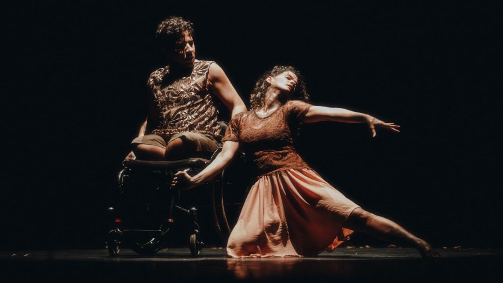 Cena de dança: um bailarino em cadeira de rodas e uma bailarina ajoelhada no chão interagem sob um feixe de luz em um palco escuro.