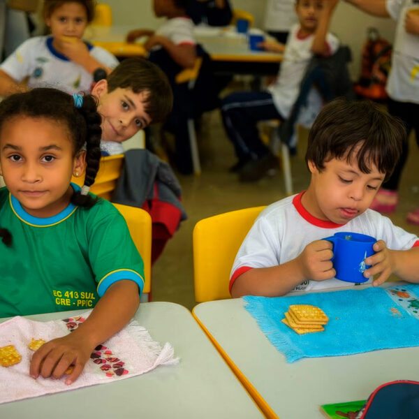 Menina negra e menino com Síndrome de Down lancham juntos sentados em suas classes numa sala de aula inclusiva.