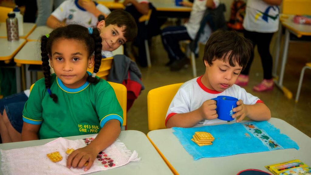 Menina negra e menino com Síndrome de Down lancham juntos sentados em suas classes numa sala de aula inclusiva.