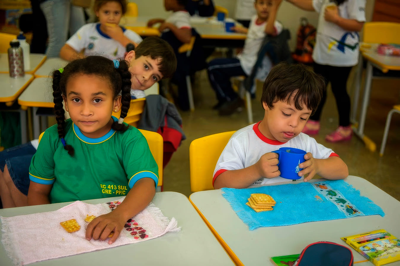 Menina negra e menino com Síndrome de Down lancham juntos sentados em suas classes numa sala de aula inclusiva.