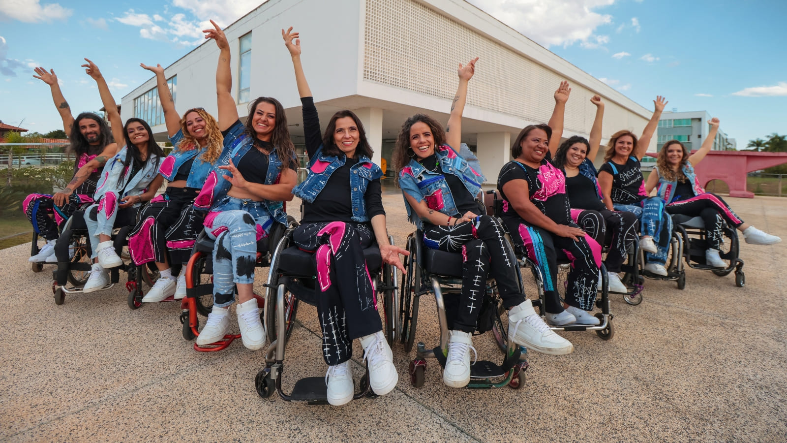 Grupo com dez integrantes do Street Cadeirante posa ao ar livre. Eles estão em suas cadeiras de rodas, usam roupas pretas e jeans com detalhes rosa e erguem os braços sorrindo.
