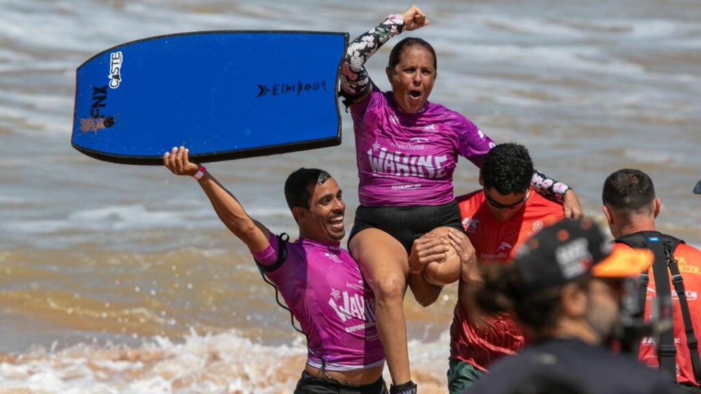 Atleta amputada comemora vitória sendo carregada nos ombros por dois homens na praia, com uma prancha azul erguida.