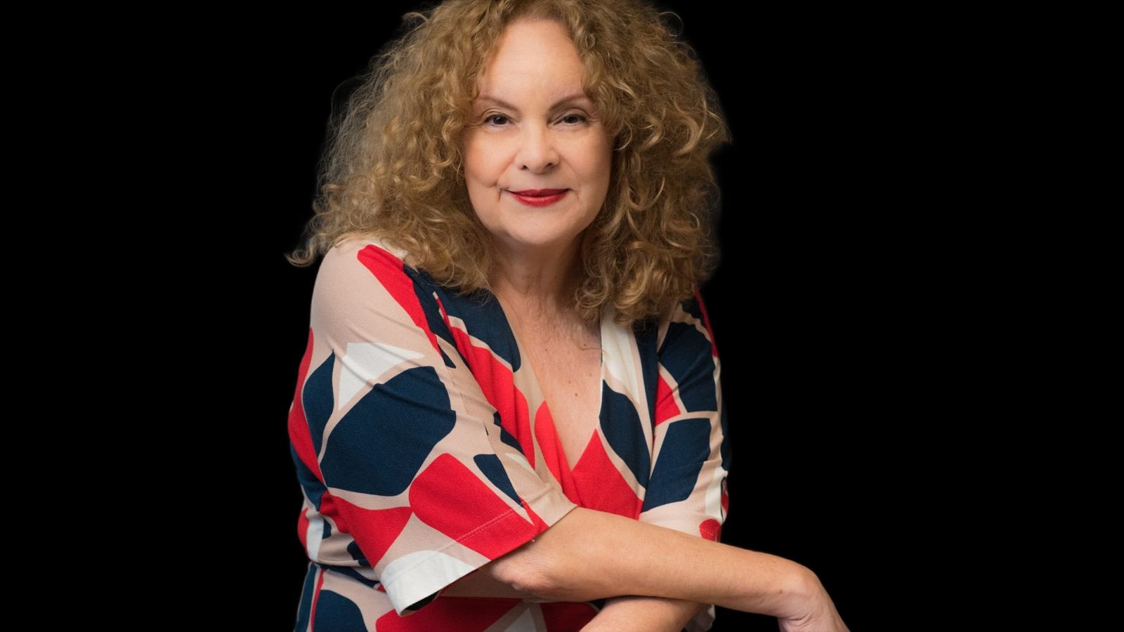 Silvana Louro, de cabelos loiros cacheados e blusa estampada em vermelho, azul e bege, posa com braços apoiados sobre fundo preto.