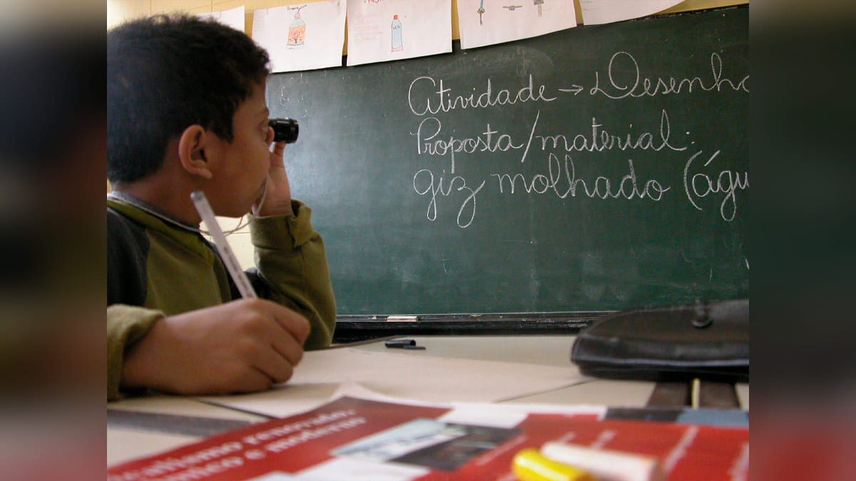 Criança com deficiência visual em sala de aula, usando uma telelupa monocular. Em sua mesa se vê algum material didático em braille.