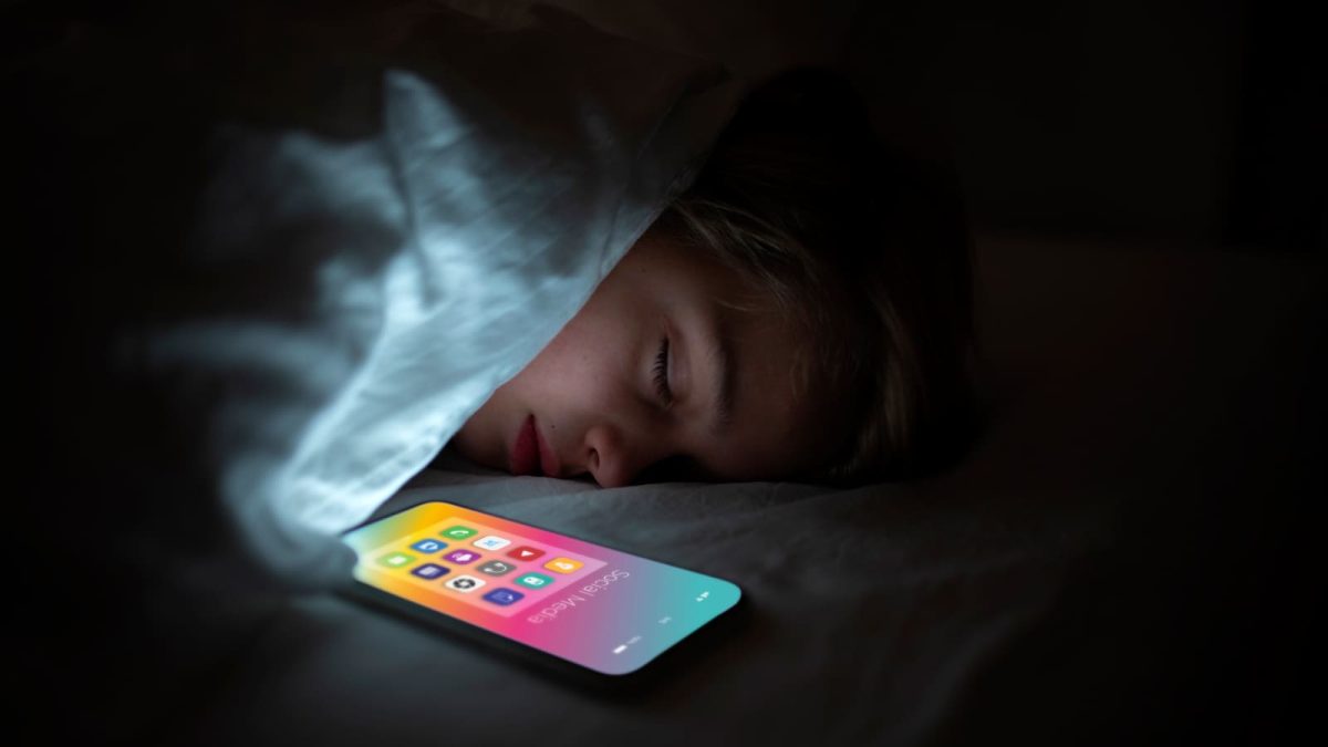 Criança dormindo com smartphone ao lado do travesseiro. Ilustra o Setembro Amarelo, sobre os riscos do vício em tecnologia para a saúde mental.